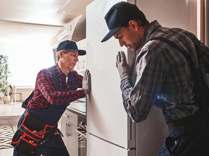 Two men in plaid shirts and hats are working on a refrigerator.